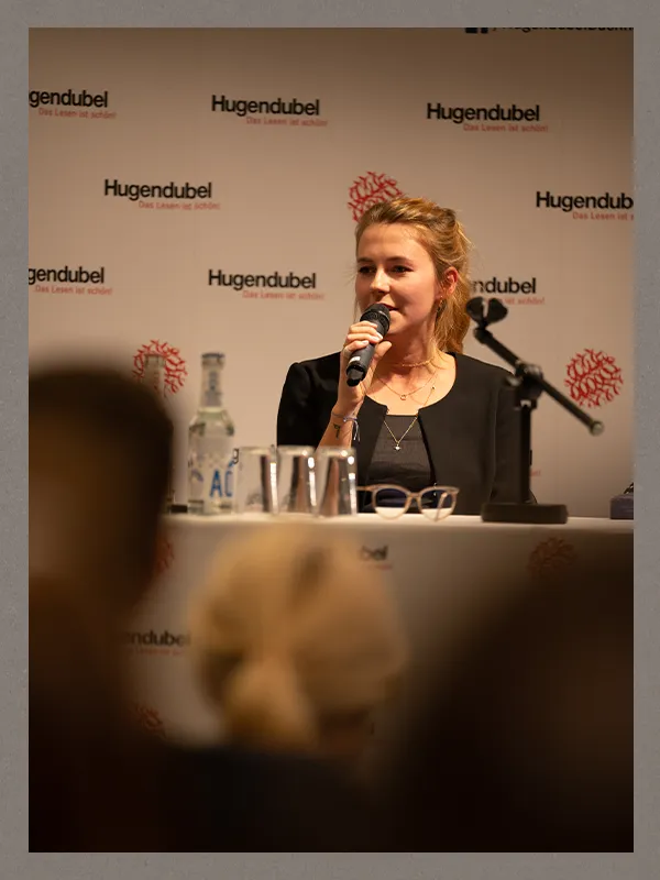 Alexandra Flint liest bei einer Lesung mit Mikrofon, im Hintergrund das Hugendubel-Logo